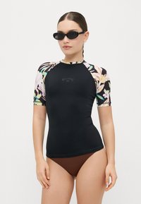 Rash guard negro con mangas cortas, acentos con patrón floral y detalle del logo en el pecho. Usado con un bikini marrón y gafas de sol.
