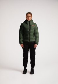 Grønne pufferjakke med høj krave, lynlåslommer og et slankt, quilted design; båret med sorte bukser og sportssko.