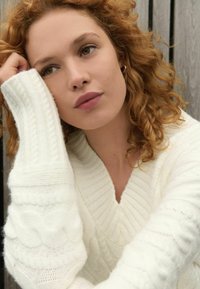 Pulover alb tricotat cu decolteu în V și modele texturate. Modelul are părul creț și o expresie neutră, cu mâna odihnită pe bărbie.