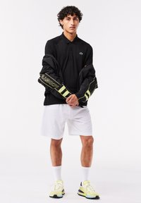 Černé polo tričko se zeleným logem, spárované s bílými šortkami. Černá bunda se žlutými pruhy přehozená přes ramena a tenisky s neonovými akcenty.