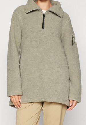 Pull polaire gris clair avec col à zip jusqu’au quart et détail d'écusson sur la manche gauche, porté avec un pantalon beige.