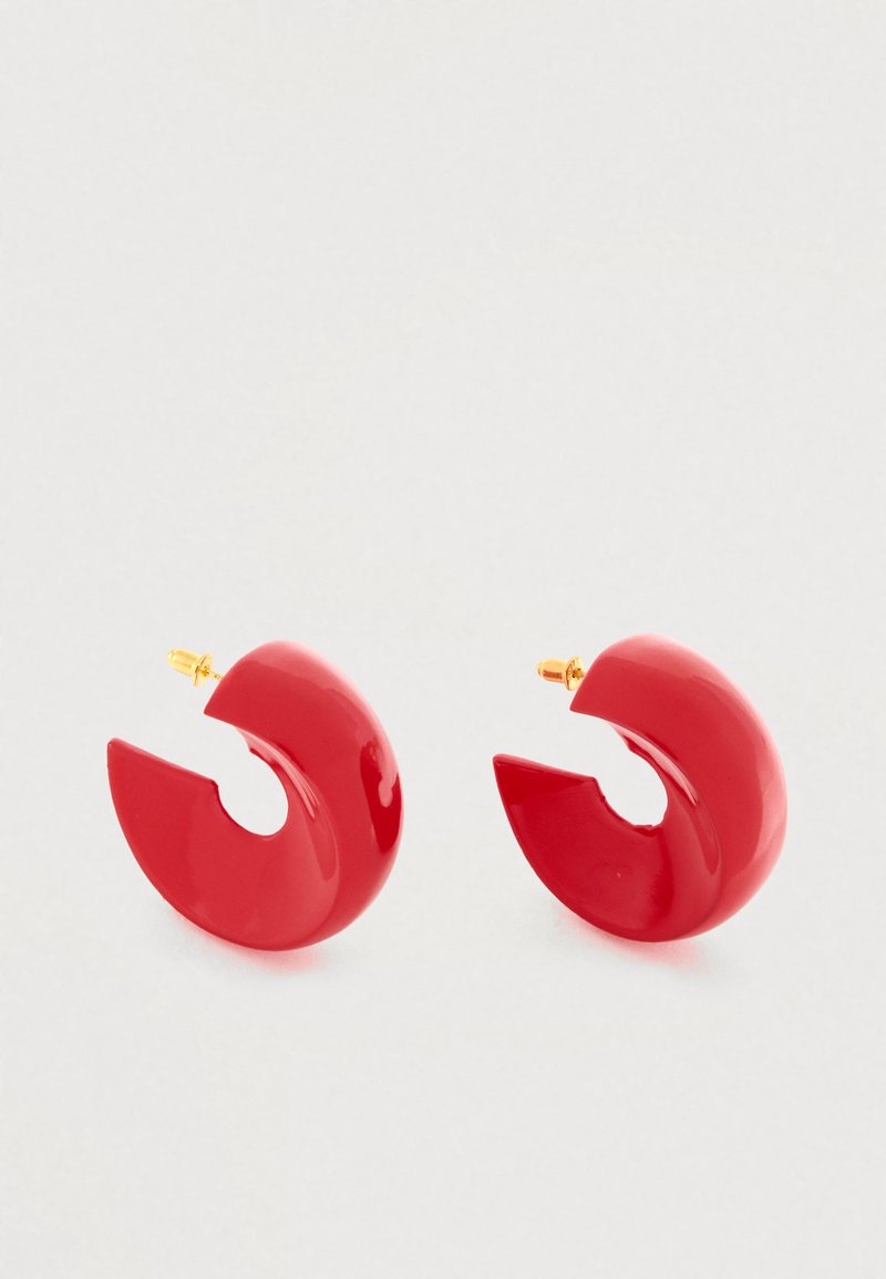 Czerwone plastikowe kolczyki w kształcie koła z połyskującym wykończeniem, posiadające unikalny otwarty wzór oraz złocone zapięcie.