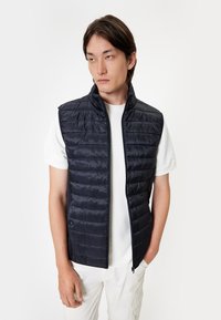 Navy blue padded vest with a high collar, zip closure, and horizontal quilted design; worn over a white t-shirt. Smooth, shiny texture.