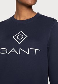 Osoba nosząca granatowy sweter z haftowanym białym logo GANT na piersi, skrócony przy szyi i ramionach.