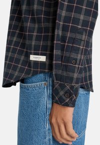 Langærmet ternet skjorte i marineblå, rød og hvide nuancer. Har knapversioner ved ærmerne, en label med teksten "ELEMENT CO.", og en afslappet pasform. Sammen med blå jeans.