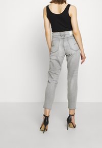 Jeans de tiro alto en gris claro con piernas ajustadas, que presentan costuras visibles y dos bolsillos traseros. Combinados con tacones altos negros con tiras.