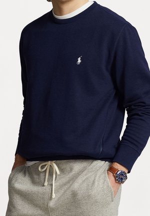 Mężczyzna ubrany w granatową bluzę z logo białego gracza polo, szare spodnie dresowe ze ściągaczem oraz brązowy skórzany zegarek z niebieską tarczą.