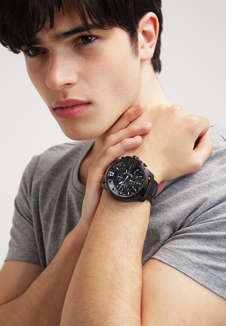 Junger Mann in grauem T-Shirt, der eine große schwarze Metallarmbanduhr trägt, hält sein Handgelenk nahe am Hals und schaut leicht zur Seite.