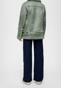 Persoon in een groene sheepskin pilotenjas, donkere wijde jeans en off-white sneakers, staand tegen een effen lichte achtergrond.