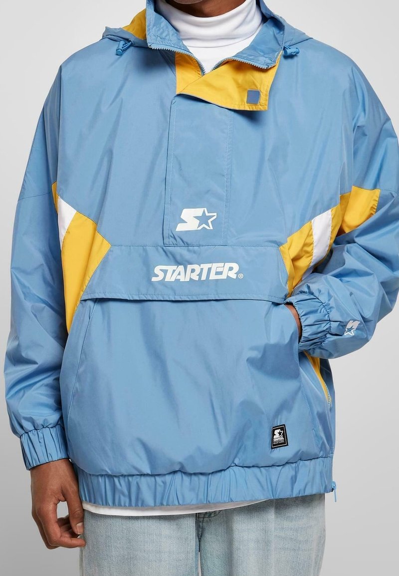 Giacca con cappuccio di un azzurro chiaro con accenti gialli e bianchi. Polsini elasticizzati, cappuccio regolabile e grande logo sul frontale. Tessuto liscio.