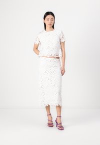 Completo bianco in due pezzi composto da un top corto e una gonna al ginocchio realizzati in pizzo floreale. Indossato con sandali con tacco alto viola.