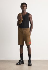 Bărbatul stă sprijinit de un perete simplu, purtând un top negru fără mâneci, pantaloni scurți maro cu șnur, șosete negre și pantofi negri, ținând o piesă vestimentară maro pe umăr.