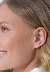 Boucles d'oreilles en forme d'étoile en métal doré rose, présentant un design ouvert. La boucle d'oreille est portée sur l'oreille d'un modèle, mettant en valeur son apparence délicate.