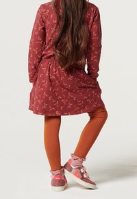 Enfant portant une robe fleurie couleur rouille, des leggings assortis, des chaussettes roses à volants et des baskets roses, debout, dos tourné.