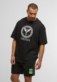 Mann mit kurzen Haaren, der ein schwarzes Colucci-T-Shirt mit dem Logo einer geflügelten Figur und schwarze Shorts mit einem Patch des Fussballclubs Kitzbühel trägt.