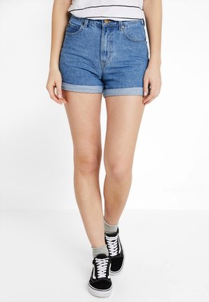 Shorts en jean de couleur bleu clair, taille haute avec des ourlets retroussés. Portés avec des baskets noires et des chaussettes grises, dotés de poches avant et d'une fermeture à boutons.
