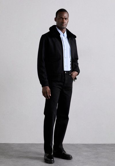 Svart ulljacka med pälskrage, ljusblå skjorta med knappar, svarta jeans och svarta skor. Minimalistisk design med rena linjer.