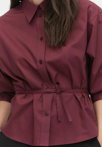 Bordeaux knap-shirt med krave og korte ærmer, med en knytning i taljen og en glat, let skinnende overflade.