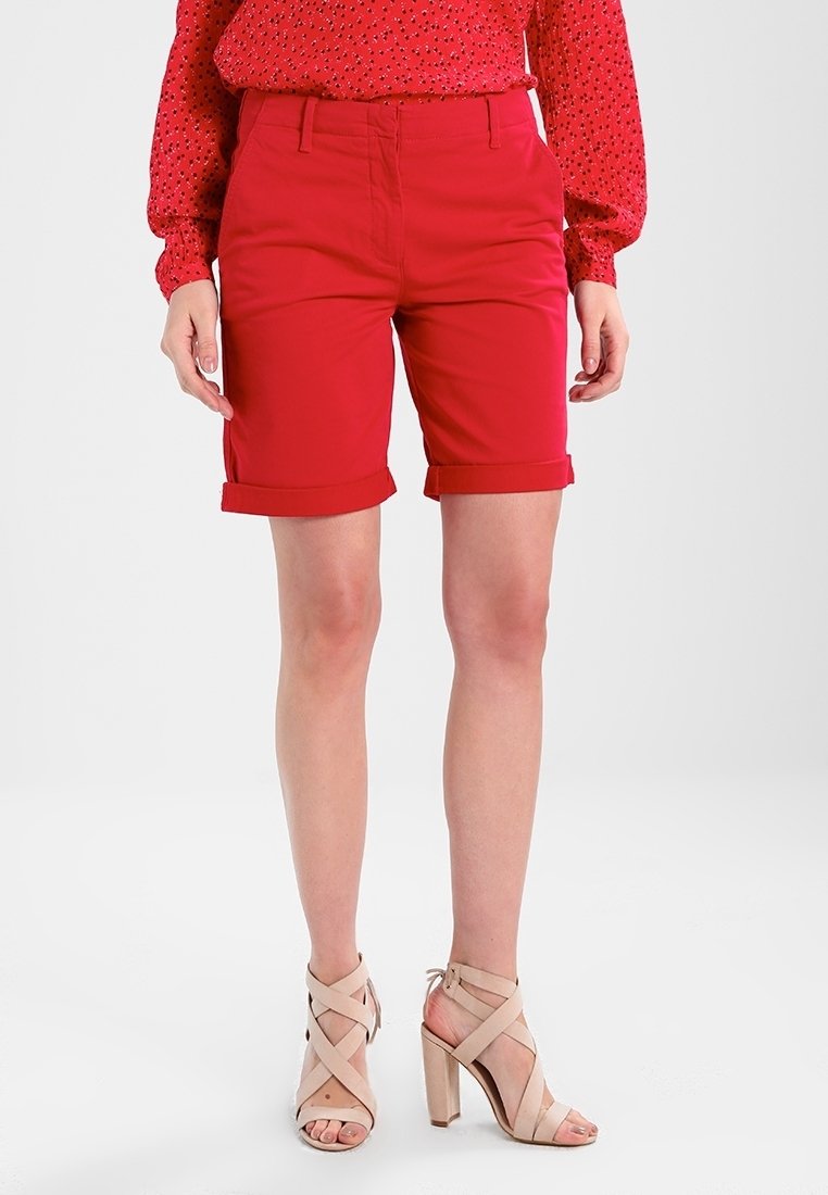 Femme portant un short rouge jusqu’aux genoux, un chemisier rouge à manches longues avec de petits motifs noirs, et des sandales à talons hauts beiges.