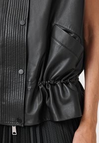 Black leather vest featuring a textured panel, gathered waist, side pocket, and round snaps. Accentuated with smooth and pleated fabric details.