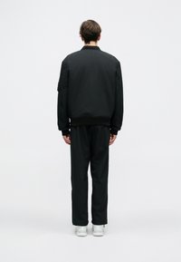 Melna bumbieru jakas ar ribotām piedurknēm un jostu, uzvilkta virs melniem biksēm. Modelis ir pagriezies ar muguru, izceļot brīvo piegriezumu un gludo tekstūru.