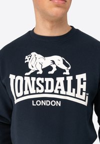 Navy sweatshirt made of cotton, featuring a white lion graphic and "LONSDALE LONDON" text in bold. Ribbed crew neckline and cuffs.