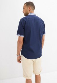 Camicia a maniche corte blu navy con colletto e polsini a quadri, abbinata a pantaloni corti beige arrotolati. Materiale in cotone con una texture liscia.