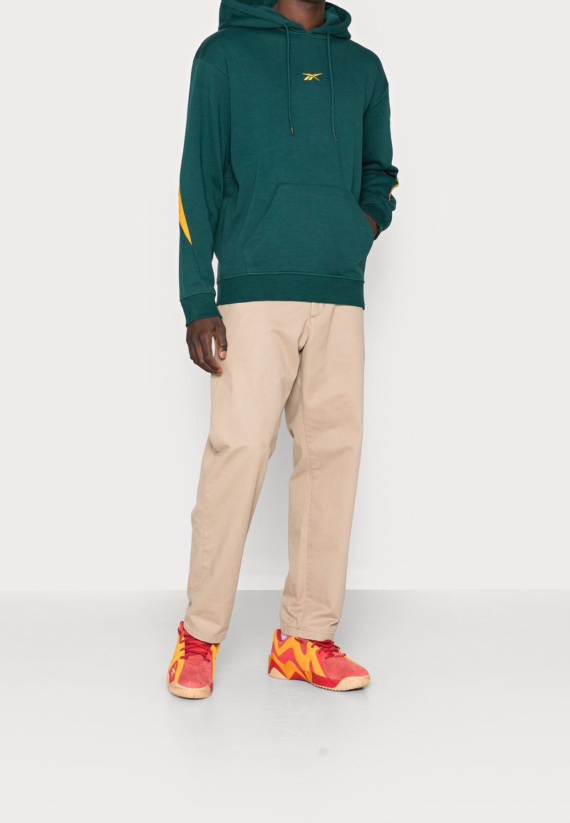 Homme debout portant un hoodie vert foncé à détails jaunes, un pantalon beige et des baskets de sport rouge-orangé sur fond uni.