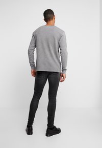 Camisa gris de manga larga con cuello redondo, combinada con jeans negros ajustados y zapatos negros, vista desde la parte posterior sobre un fondo liso.