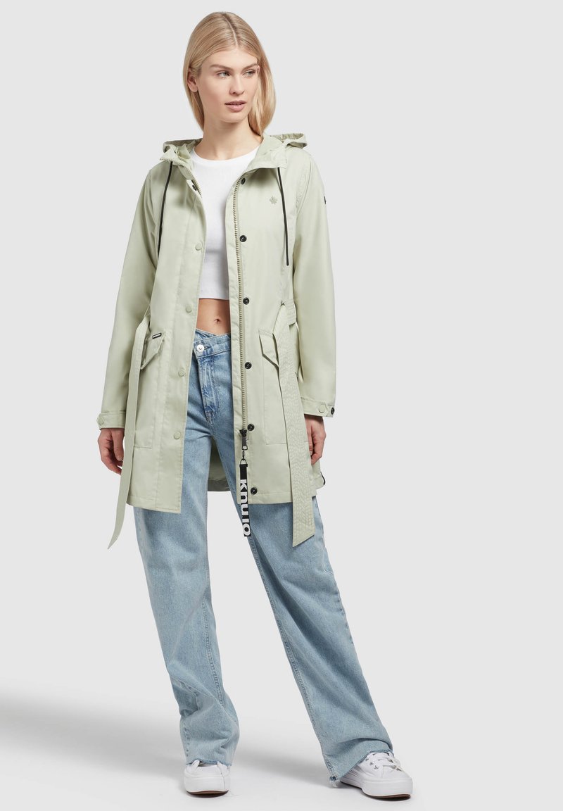 Giacca impermeabile verde chiaro con cappuccio, chiusure a bottone, lacci in vita e due tasche frontali. Abbinata a jeans a gamba larga azzurri.