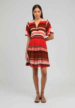 Femme debout portant une robe rayée rouge, rose et marron mi-longue, avec des manches courtes et des sandales ouvertes, sur un fond uni.