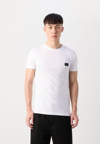 T-shirt bianco a maniche corte realizzato in tessuto morbido, con un piccolo dettaglio di tasca nera sul lato sinistro del petto. Design minimalista, forma attillata.