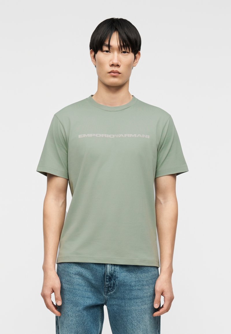 Άνδρας με μαύρα μαλλιά που φοράει ανοιχτοπράσινη μπλούζα Emporio Armani και μπλε τζιν, στέκεται μπροστά σε ένα απλό φωτεινό φόντο.