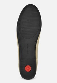 Gumowa podeszwa o teksturowanym czarnym wykończeniu, z czerwonym ośmiokątnym akcentem i wytłoczonym logo. Górna część z kompozytu w jasnym odcieniu brązu.