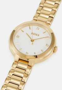 Montre bracelet dorée avec un boîtier rond, un cadran blanc, des index dorés et trois petits diamants. Elle est dotée d'un bracelet en maillons métalliques.