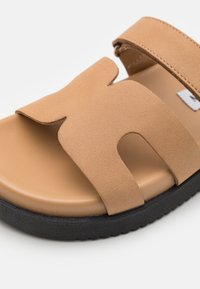 Tan syntetisk halkande sandal med en bred remdesign med utskärningar, en slät fotbädd och en strukturerad svart gummisula.