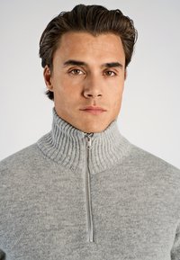 Grå zip-up tröja med ribbad hög krage och mjuk textur, med en silverfärgad dragkedja framtill. Ren, minimalistisk design.