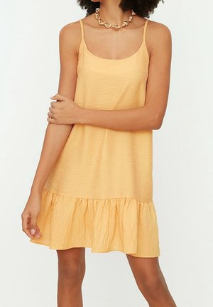 Kobieta w żółtej sukience bez rękawów z falbanką na dole i masywnym złotym naszyjnikiem z koralików, stojąca z założonymi ramionami.