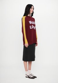 Burgundy knit sweater with knitted “MAKE LOVE” text, contrasting yellow sleeves, worn over a black skirt with flat shoes and white socks.