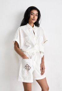 Λευκό 100% βαμβακερό κοστούμι με γιακά, πλαϊνές τσέπες με δαντέλα και waist με κορδόνι για ρυθμιζόμενη εφαρμογή.