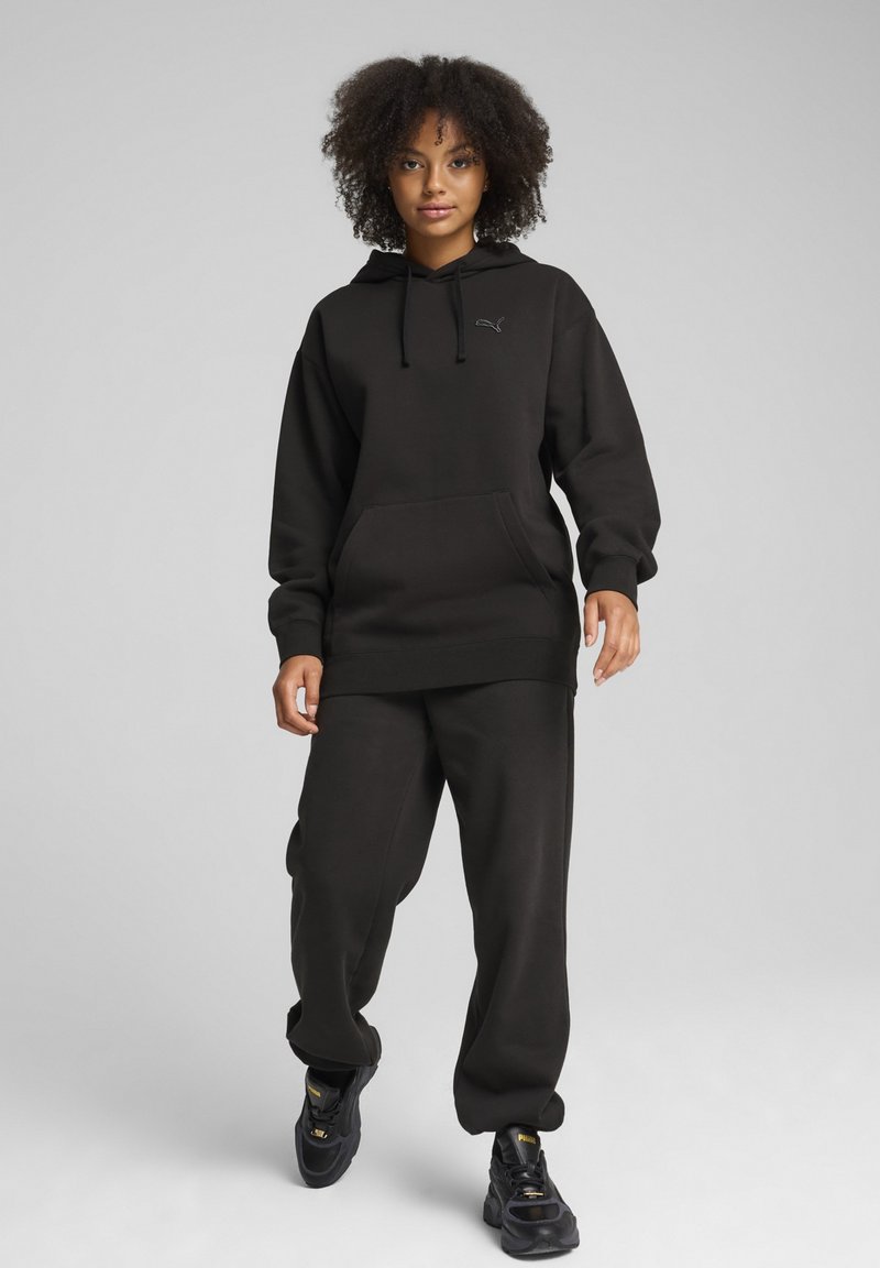 Sort hættetrøje og matchende sweatpants, lavet af blødt stof. Hættetrøjen har snore og en frontlomme. Bæres med sorte træningssko.
