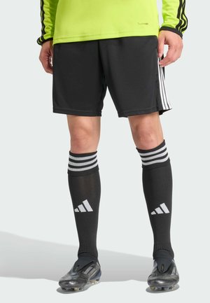Mann trägt schwarze Adidas Fußballshorts, schwarze Adidas knielange Socken, schwarze Stollenschuhe und ein neongelbes Langarmshirt und steht.
