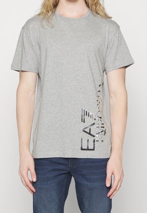 Osoba nosząca szarą koszulkę z pionowym czarnym logo EA7 Emporio Armani po prawej stronie, połączoną z niebieskimi jeansami.