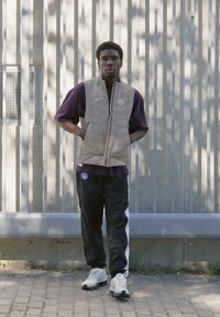 Puff vest in light beige, purple short-sleeve shirt, black track pants with white stripes, and white sneakers. Standing against a textured wall.