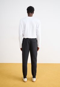 Λευκό πουκάμισο με μακριά μανίκια, μαύρο παντελόνι με λεπτές ρίγες και λευκά αθλητικά παπούτσια με το λογότυπο "Nike", εμφανιζόμενο από πίσω σε μια απλή φόντο.