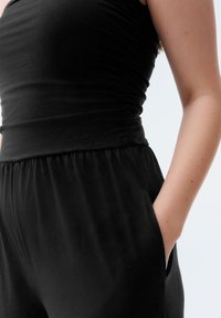 Black jumpsuit featuring a fitted bodice, gathered texture, wide waistband, and side pockets. Soft fabric with a smooth finish.