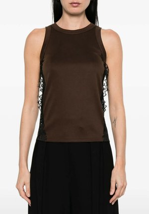 Donna che indossa un top marrone senza maniche con pannelli laterali in pizzo nero e pantaloni neri, in piedi con le braccia rilassate lungo i fianchi.