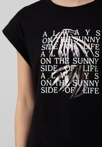 Zwarte katoenen T-shirt met opgerolde mouwen, voorzien van witte en lichtgekleurde tekst en een bladvormig grafisch ontwerp dat op de voorkant is gedrukt.