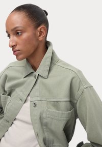 Giacca di denim verde chiaro con colletto, tasca sul petto e bottoni a pressione argentati. Presenta una vestibilità rilassata e dettagli di cucitura ben visibili.