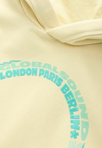 Gelbe Kapuzenjacke mit einem türkisfarbenen, kreisförmigen Grafikdesign und dem Text "GLOBAL SOUND LONDON PARIS BERLIN." Weicher Stoff mit lässigem Design.